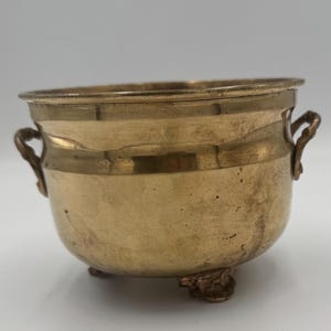 May include: A small, antique brass bowl with two handles and three decorative feet. The bowl has a rounded shape and a polished finish, with a slight tarnish adding character. The handles are curved, and the feet have a detailed, ornate design.