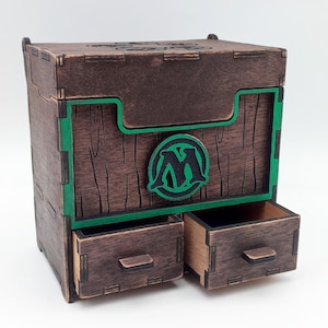 May include: A wooden box with two drawers, stained in a dark brown color. The box features green accents and a circular emblem with a stylized "M" in green and black. The drawers are open, revealing the interior.