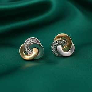 May include: A pair of stud earrings with a three-loop design. Each earring features a gold-colored loop, a silver-colored loop, and a loop encrusted with small, clear gemstones. The earrings are set against a dark green, silky background.
