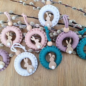 Op de afbeelding: Een verzameling paasornamenten. De afbeelding toont macramékransen in wit, roze, teal en paars, elk met een houten konijn en een houten kraal om op te hangen. De kransen worden weergegeven op een houten oppervlak met wilgentakken op de achtergrond.