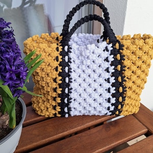 Peut inclure: Un sac fourre-tout macramé fait à la main avec un motif géométrique. Le sac présente une combinaison de jaune moutarde, de blanc et de noir. Les poignées sont noires. Le sac est posé sur une surface en bois à côté d'une fleur violette en pot.