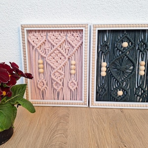 May include: Two framed macrame wall hangings. One is light pink with wooden beads, the other is dark teal. Both have white frames with wooden bead trim. A potted plant with dark red flowers is in the foreground.