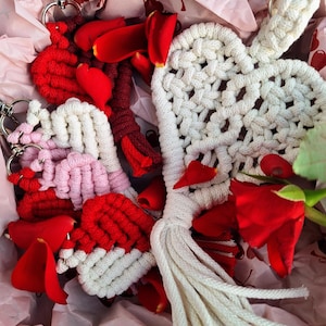 May include: A collection of handmade macrame heart keychains in red, white, and pink, with silver key rings. The keychains are nestled in a box with red rose petals and pink tissue paper. A white macrame heart with a tassel is also visible.