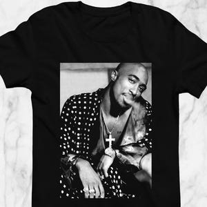 May include: Black t-shirt featuring a black and white image of a person wearing a polka dot jacket, a cross pendant, and rings. The image is a portrait of a famous rapper.