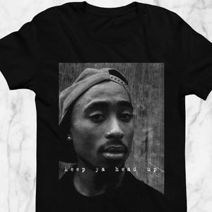 May include: Black t-shirt featuring a black and white portrait of a person wearing a baseball cap. The image includes the text "keep ya head up" in white font. The shirt is displayed on a marble surface.