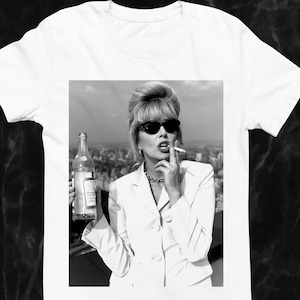 May include: White t-shirt with a black and white image of a woman in sunglasses smoking a cigarette and holding a bottle. The woman is wearing a white suit and a silver necklace. The background shows a cityscape.