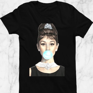 May include: Black t-shirt featuring a graphic of Audrey Hepburn blowing a bubblegum bubble. The image shows Hepburn wearing a pearl necklace, a black dress, and a tiara. The t-shirt is made of a soft, comfortable material.