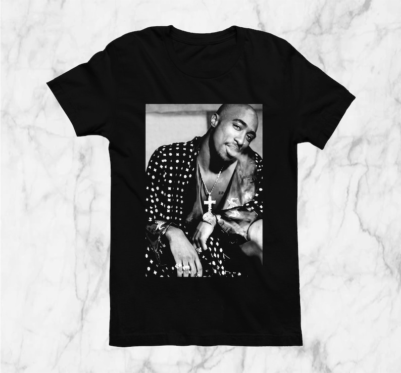 May include: Black t-shirt featuring a black and white image of a person wearing a polka dot jacket, a cross pendant, and rings. The image is a portrait of a famous rapper.