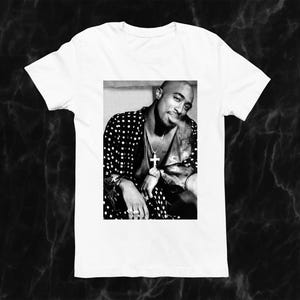 May include: White t-shirt featuring a black and white image of a person wearing a patterned jacket and cross pendant. The image is a portrait of a person with a slight smile.