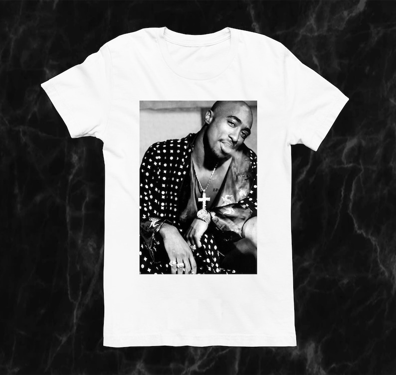 May include: White t-shirt featuring a black and white image of a person wearing a patterned jacket and cross pendant. The image is a portrait of a person with a slight smile.