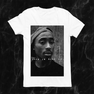 May include: White t-shirt featuring a black and white image of a person wearing a cap. The image includes the text "keep ya head up" in a stylized font. The shirt is a classic crew neck style.