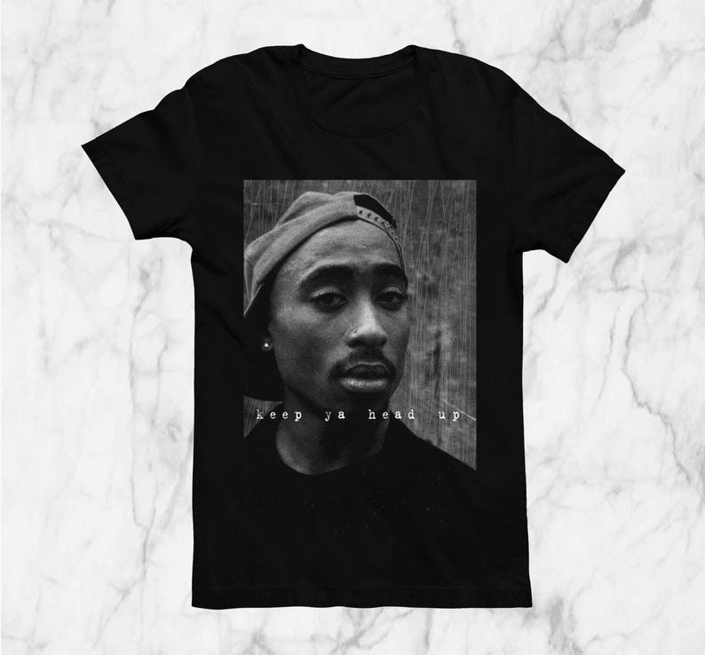 May include: Black t-shirt featuring a black and white portrait of a person wearing a baseball cap. The image includes the text "keep ya head up" in white font. The shirt is displayed on a marble surface.