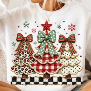 May include: A white crewneck sweatshirt featuring a graphic of three Christmas trees. The left tree has a leopard print, the center tree has a red and white buffalo plaid pattern, and the right tree has a white polka dot pattern. Each tree is topped with a large bow and a red star.