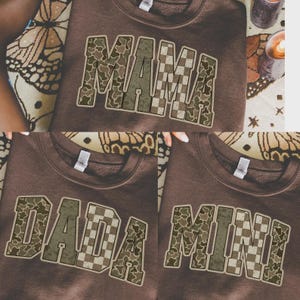 May include: A dark brown crewneck sweatshirt featuring the word MAMA in large block letters. The letters have a mixed pattern of green camouflage and a tan checkered print. The design is repeated on other sweatshirts with the words DADA and MINI.