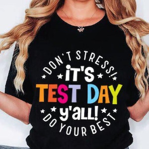 Peut inclure: T-shirt noir avec le texte "DON'T STRESS! IT'S TEST DAY y'all! DO YOUR BEST" dans un design circulaire color&eacute;. Le t-shirt est &agrave; col rond et &agrave; manches courtes.
