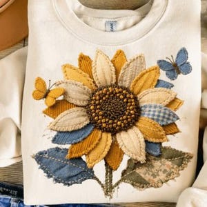 May include: Cream-colored sweatshirt featuring a large, appliqued sunflower with yellow, tan, and blue fabric petals. Two butterfly appliqués are also present. The sunflower design is centered on the front of the shirt.