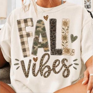 May include: An off-white short-sleeve t-shirt featuring the text FALL VIBES. The word FALL is printed in block letters with plaid, camouflage, leopard, and sunflower patterns. The word VIBES is in cursive with a leopard print and small hearts.