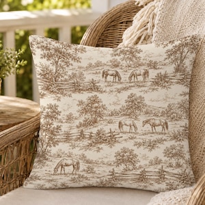 May include: A square throw pillow featuring a cream-colored fabric with a brown toile-style print. The pattern depicts a pastoral landscape with multiple horses grazing in fields, scattered trees, and wooden fence lines across the surface.