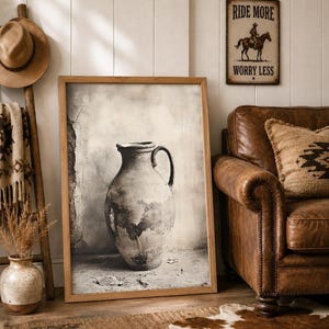 May include: A framed wall art print featuring a monochromatic image of a weathered ceramic pitcher with a single handle. The artwork is displayed in a light wood frame, showcasing a textured, aged aesthetic with neutral gray and beige tones.