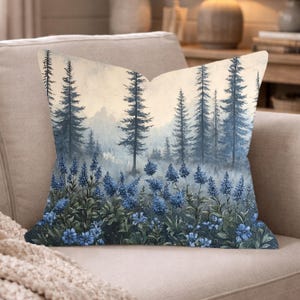 May include: A square throw pillow featuring a printed landscape scene of tall, dark blue pine trees set against a misty, light-colored background. The foreground is filled with clusters of vibrant blue flowers and green foliage.