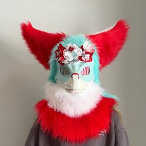 以下が含まれることがあります： 赤と青のアクセントが施されたカラフルな毛皮のマスク。 マスクは大きな赤い耳、水色の顔、白と赤の毛皮の襟が特徴です。 花の冠がマスクの上部を飾っています。