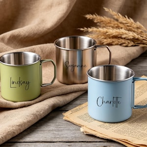 May include: Three personalized metal camping mugs in green, silver, and blue. Each mug has a silver rim and handle, with a name printed in black script: "Lindsay", "Suzanne", and "Charlotte". The mugs are on a wooden surface.