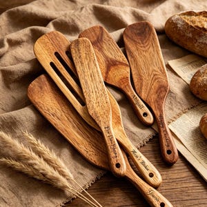 May include: A collection of wooden kitchen utensils including spatulas and a slotted turner with engraved text. The utensils feature natural wood grain patterns and are arranged alongside three crusty loaves of bread and dried wheat stalks.