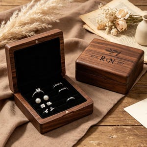 May include: Two dark brown wooden ring boxes with black velvet interiors. One box is open, displaying a diamond ring, pearl earrings, and small silver studs. The closed box features a laser-engraved branch design, the letters R and N, and 02.14.2027.