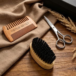 May include: A beard grooming set consisting of a light brown wooden comb with the text DAD'S BEARD KIT engraved on it, a small oval wooden brush with black bristles, and a pair of silver metal grooming scissors, all arranged together.