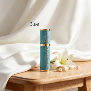 K&ouml;nnte beinhalten: Eine zylindrische Parf&uuml;mflasche in T&uuml;rkisblau mit goldenen Akzenten. Die Flasche hat eine strukturierte, lederartige Oberfl&auml;che und tr&auml;gt die Aufschrift "Blue". Eine wei&szlig;e Lilie und Perlen liegen auf einer Holzoberfl&auml;che.