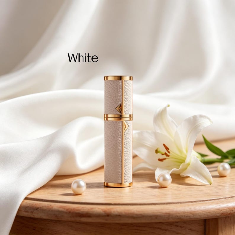 K&ouml;nnte beinhalten: Ein wei&szlig;er Parf&uuml;mzerst&auml;uber mit goldenen Akzenten. Der Zerst&auml;uber hat eine strukturierte, lederartige Optik und ist mit "White" beschriftet. Eine wei&szlig;e Lilienbl&uuml;te und Perlen sind auf einer Holzoberfl&auml;che angeordnet.