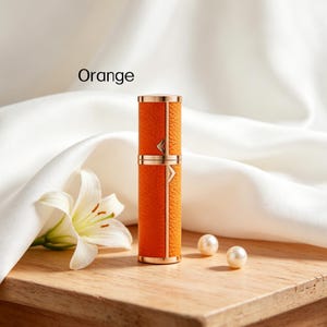 K&ouml;nnte beinhalten: Ein orangefarbener und goldfarbener Parf&uuml;mzerst&auml;uber mit dem Wort "Orange" im Bild. Der Zerst&auml;uber hat eine lederartige Textur und goldfarbene Akzente. Zwei Perlen und eine wei&szlig;e Blume sind ebenfalls im Bild.