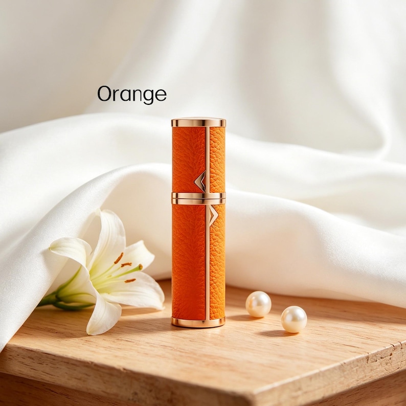 K&ouml;nnte beinhalten: Ein orangefarbener und goldfarbener Parf&uuml;mzerst&auml;uber mit dem Wort "Orange" im Bild. Der Zerst&auml;uber hat eine lederartige Textur und goldfarbene Akzente. Zwei Perlen und eine wei&szlig;e Blume sind ebenfalls im Bild.