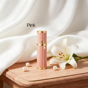 K&ouml;nnte beinhalten: Rosa Parf&uuml;mzerst&auml;uber mit goldenen Akzenten. Die zylindrische Flasche hat eine strukturierte rosa Au&szlig;enseite und goldfarbene B&auml;nder und Details. Das Wort "Pink" ist auf dem Bild aufgedruckt. Eine wei&szlig;e Lilie und Perlen sind ebenfalls im Bild.