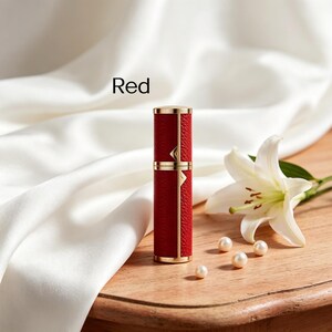 K&ouml;nnte beinhalten: Eine rote, zylindrische Parf&uuml;mflasche mit goldenen Akzenten und einer strukturierten Lederoptik. Das Wort "Red" ist auf dem Bild zu sehen. Die Flasche steht neben einer wei&szlig;en Lilie und Perlen auf einer Holzoberfl&auml;che.
