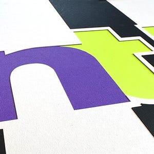 May include: Abstract composition with large, layered letters in purple, white, black, and lime green. The letters are cut out and arranged to create a dynamic, graphic design. The image has a modern, artistic feel.
