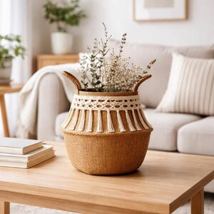 May include: A brown woven basket with a cream-coloured crocheted band and tassels, filled with dried flowers. The basket has two handles and sits on a wooden table. The basket's base is round.