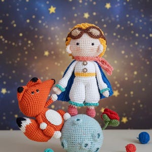 Puede incluir: Figuras de ganchillo de un niño con capa, un zorro, una rosa y un pequeño planeta. El niño lleva un traje blanco, gafas y una bufanda. El zorro es naranja y blanco. El fondo es una noche estrellada.