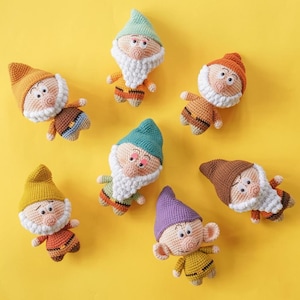 以下が含まれることがあります： さまざまな色の帽子と衣装を着た、手作りの編み人形のドワーフが7体、黄色の背景に配置されています。 各人形は白いひげ、バラ色の頬、ユニークな衣装を持っています。 人形は糸で作られています。