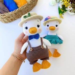 Könnte beinhalten: Zwei handgefertigte Häkelenten-Puppen. Eine Ente trägt eine braune Latzhose mit Hosenträgern, eine blaue Fliege und einen beigen Hut. Die andere Ente trägt ein türkisfarbenes Kleid und einen Hut mit floralen Akzenten. Beide haben orangefarbene Schnäbel und Füße.