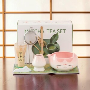 May include: A matcha tea set including a white box labelled MATCHA TEA SET, a bamboo whisk in a clear container, a metal mesh strainer, a bamboo scoop, a white whisk holder, a white mountain-shaped rest, and a pink and white cat-themed ceramic bowl.