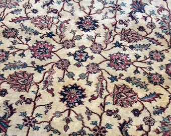 Op de afbeelding: Een rechthoekig vloerkleed met een crèmekleurige achtergrond en een ingewikkeld bloemmotief in tinten donkerrood, marineblauw en groenblauw. Het ontwerp bestaat uit onderling verbonden ranken en gestileerde bloemen, omlijst door een donkerrode rand.
