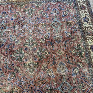 Op de afbeelding: Een rechthoekig vloerkleed met een complex bloemen- en geometrisch patroon in tinten oudroze, gedempt blauw, saliegroen en cr&egrave;me. Het ontwerp bevat een brede decoratieve rand met ingewikkelde botanische motieven rondom het centrale veld.