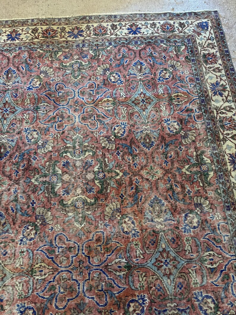 Op de afbeelding: Een rechthoekig vloerkleed met een complex bloemen- en geometrisch patroon in tinten oudroze, gedempt blauw, saliegroen en cr&egrave;me. Het ontwerp bevat een brede decoratieve rand met ingewikkelde botanische motieven rondom het centrale veld.