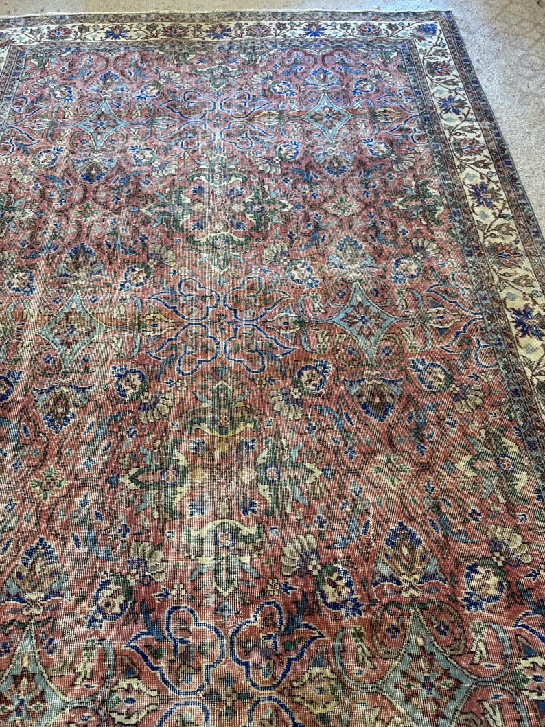 Op de afbeelding: Een rechthoekig vloerkleed met een vervaagde terracotta rode basiskleur en ingewikkelde bloem- en geometrische patronen in tinten stoffig blauw, saliegroen en cr&egrave;me. Het kleed heeft een brede decoratieve rand met herhalende bloemmotieven.