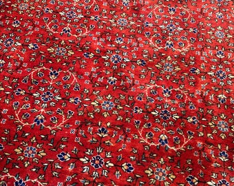 Op de afbeelding: Een rood vloerkleed met een herhalend patroon van cirkelvormige bloemenmedaillons. Het ontwerp bevat ingewikkelde ranken en kleine bloesems in tinten donkerblauw, lichtblauw en crème, wat zorgt voor een dicht, symmetrisch textieloppervlak.