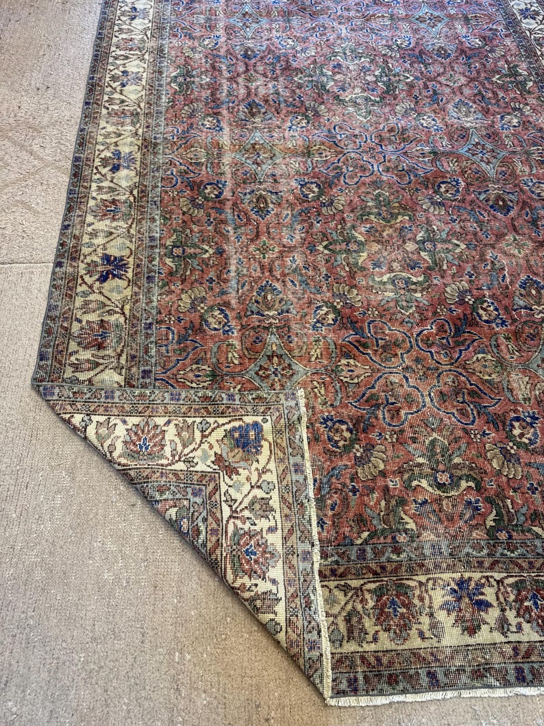 Op de afbeelding: Een rechthoekig vloerkleed met een complex bloemenpatroon in tinten terracotta, saliegroen en marineblauw. Het kleed heeft een brede cr&egrave;mekleurige rand met gedetailleerde botanische motieven en een omgeslagen hoek die de onderkant laat zien.