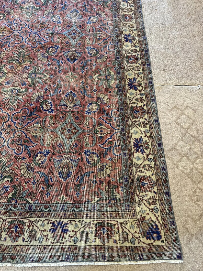 Op de afbeelding: Een rechthoekig vloerkleed met een vervaagd terracotta rood veld met ingewikkelde bloemmotieven en geometrische patronen in tinten blauw, saliegroen en cr&egrave;me. Het kleed heeft een brede, versierde rand op een cr&egrave;mekleurige achtergrond.