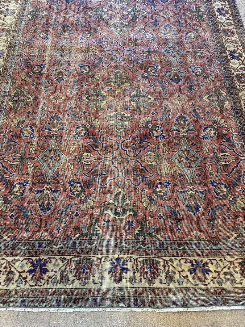 Op de afbeelding: Een rechthoekig vloerkleed met een dicht, ingewikkeld bloemen- en rankenpatroon in tinten terracotta rood, saliegroen en diepblauw. Het ontwerp bevat een brede, sierlijke rand met een cr&egrave;mekleurige achtergrond en herhalende botanische motieven.