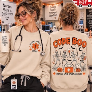 May include: A beige crewneck sweatshirt featuring a small orange medical star logo with the text Emergency Department and the name Sophia on the front, and a large graphic on the back showing four skeletons, ghosts, and the text Code Boo.